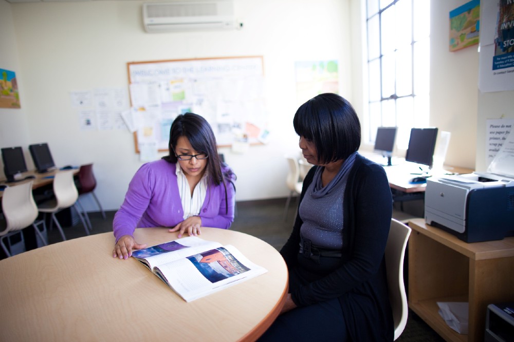 Two women seated at a table in an computer lab review a document.