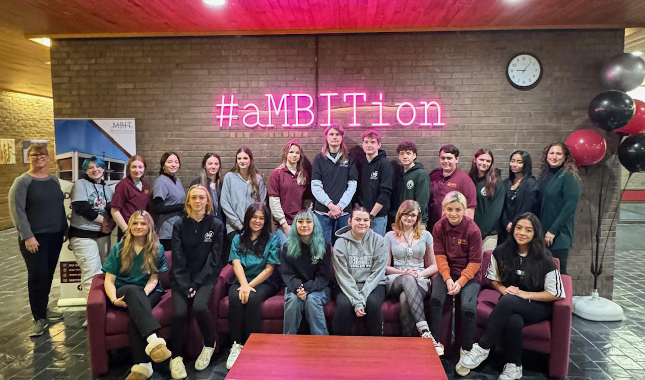 Group of students standing and sitting in front of a brick wall with a neon sign reading #ambition hanging on it